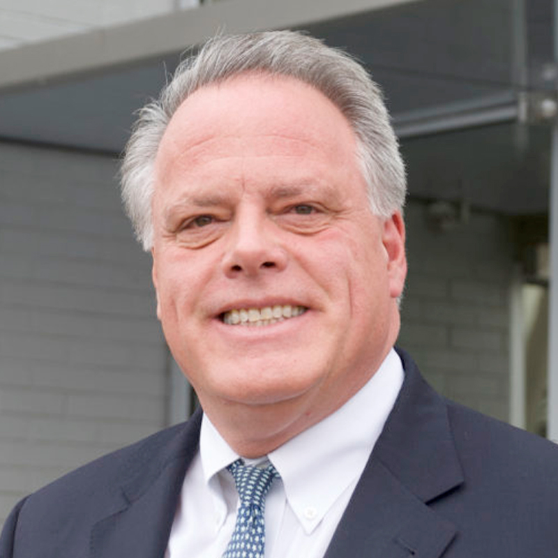 A middle-aged man with gray hair wearing a dark suit, white shirt, and patterned tie smiles outdoors in front of a modern building.