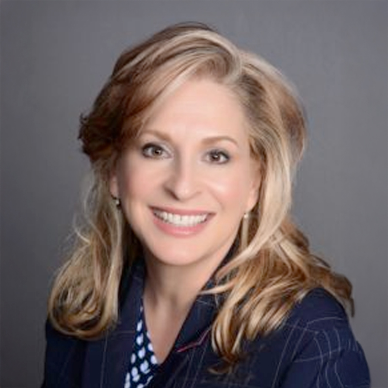 A woman with long, wavy blonde hair smiles at the camera. She is wearing a dark blazer over a patterned blouse and is posed in front of a plain gray background.