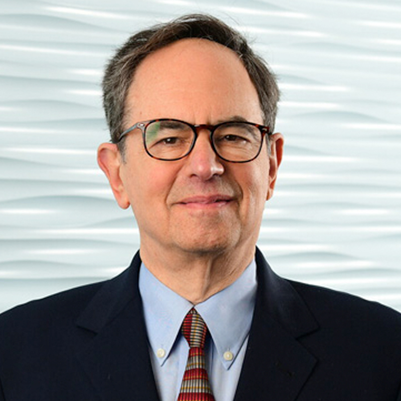 A middle-aged man with short dark hair, glasses, and a slight smile wears a dark suit, light blue shirt, and striped tie. The background features a white, wavy textured wall.