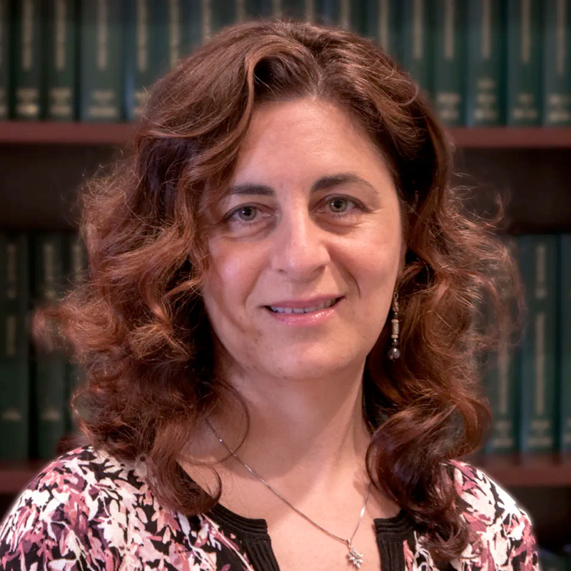 A woman with wavy brown hair, wearing a patterned top and a necklace, stands in front of a bookshelf filled with green books, smiling softly at the camera.