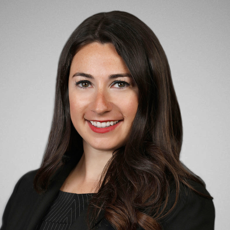A woman with long, dark brown hair smiles at the camera. She is wearing a black blazer over a dark top, and the background is a plain, light gray.