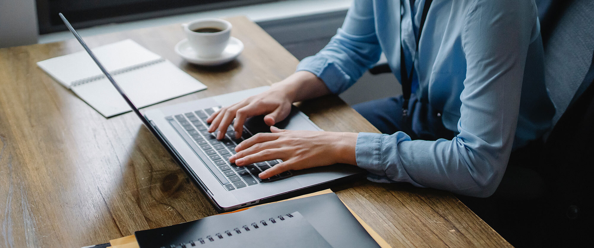A person in a blue shirt types on a laptop at a wooden desk with a notepad, pen, and a cup of coffee nearby. Natural light filters in from a window in the background, as they focus on crafting content for law firm marketing services.