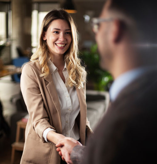 Professional young woman in beige suit with blonde hair shaking hands with professional man with brown grey suit
