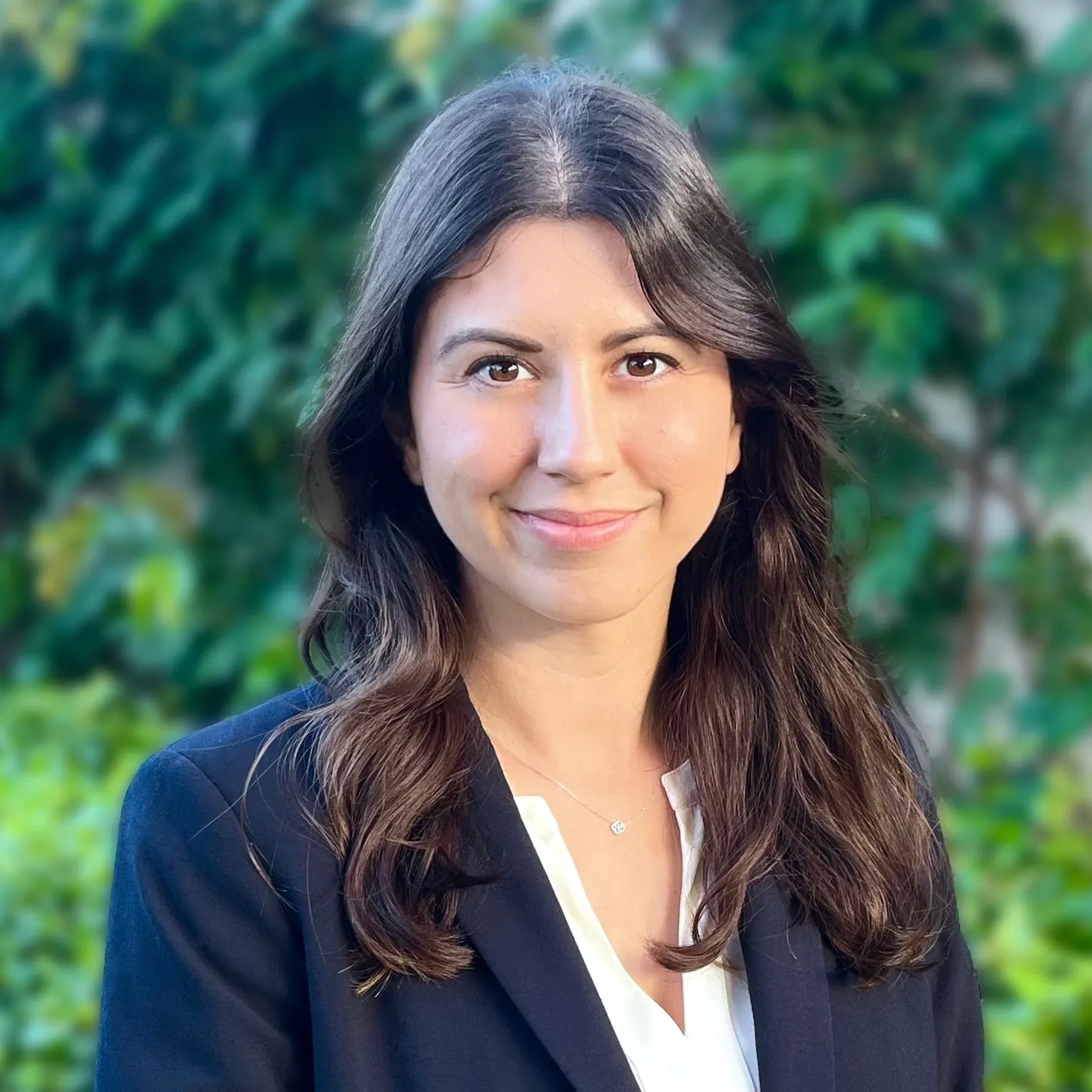 A woman with long brown hair, wearing a dark blazer over a white top, smiles softly while standing outdoors in front of green, leafy foliage.