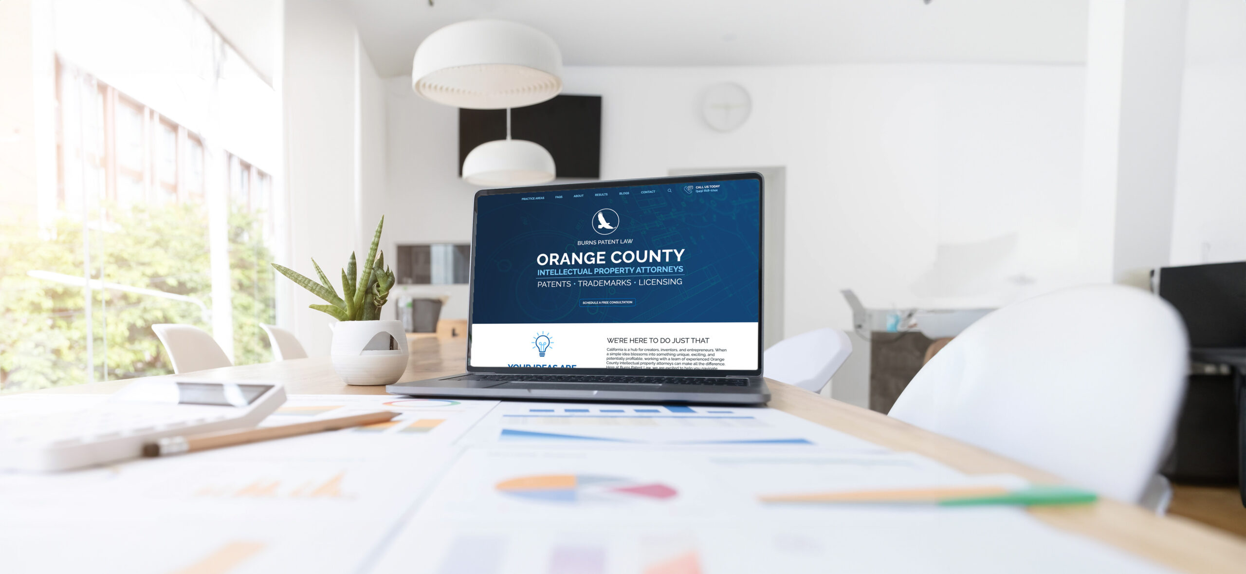 A laptop is open on a desk in a modern office space, displaying a website for Orange County law firm marketing services. The desk also has some documents with charts and graphs, a succulent plant, and cups. The office has white walls, large windows, and several white chairs.