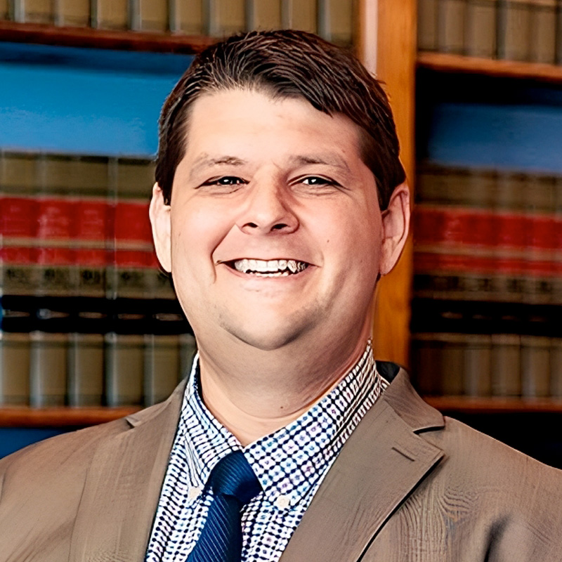 A man in a tan suit, blue checkered shirt, and blue tie smiles, standing in front of bookshelves filled with legal books.