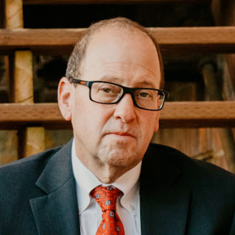 A middle-aged man wearing glasses, a dark suit, white shirt, and red patterned tie sits in front of wooden stairs, looking at the camera with a neutral expression.