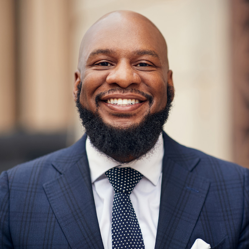 A smiling man with a bald head and full beard, wearing a blue suit, white shirt, and polka dot tie, stands outdoors with a blurred building in the background.