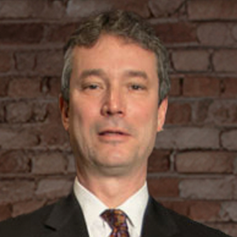 A middle-aged man with short, graying hair, wearing a dark suit, white shirt, and patterned tie, stands in front of a brick wall background.