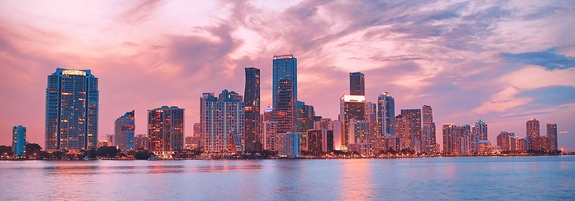 A city skyline at sunset with tall buildings reflecting on a body of water. The sky, reminiscent of Florida's vibrant evenings, is a mix of pink and purple hues, with wispy clouds creating a dramatic backdrop. Lights from the buildings start to illuminate the scene.