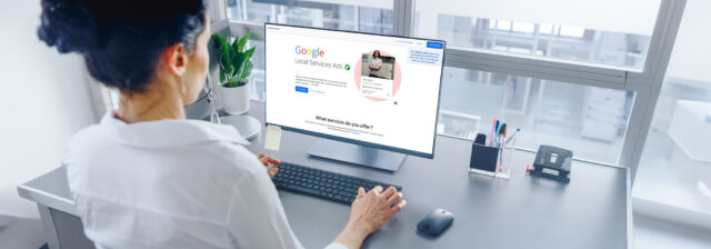 A person is sitting at a desk, looking at a computer screen displaying a Google Local Services Ads webpage, ensuring the listings are Google Screened. The workspace is tidy with a potted plant, a pen holder, and an office phone. The window in front of the person shows a blurred office environment beyond.