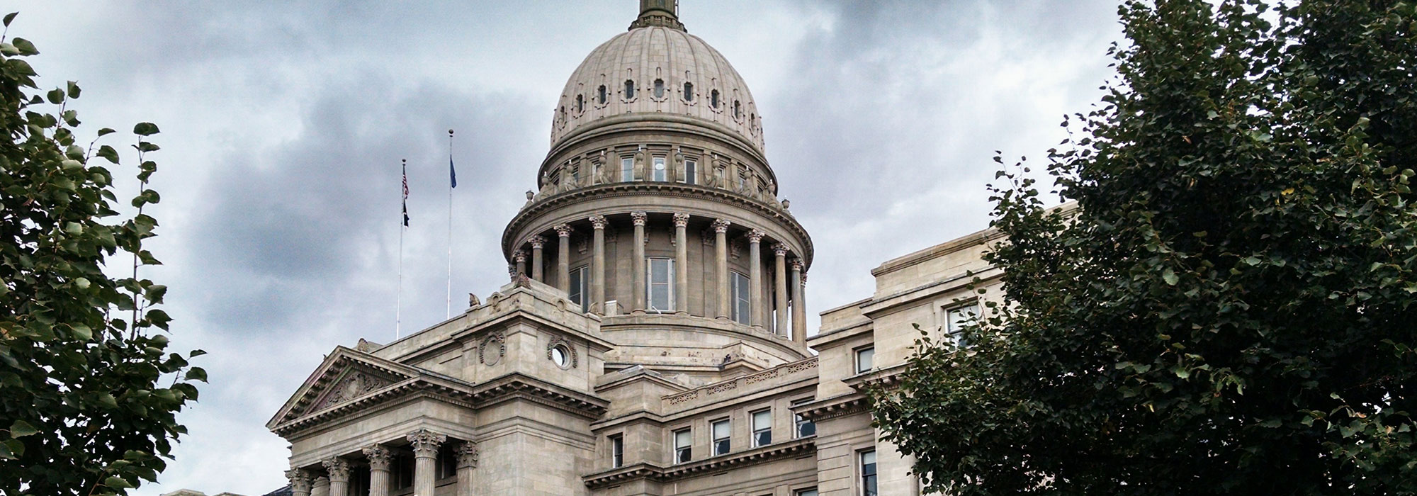 A grand historical building with a large dome and stone facade set against a cloudy sky, reminiscent of the stately courthouses governed by Idaho Attorney Advertising Rules. Trees add greenery to the scene while flags flutter atop the impressive structure.
