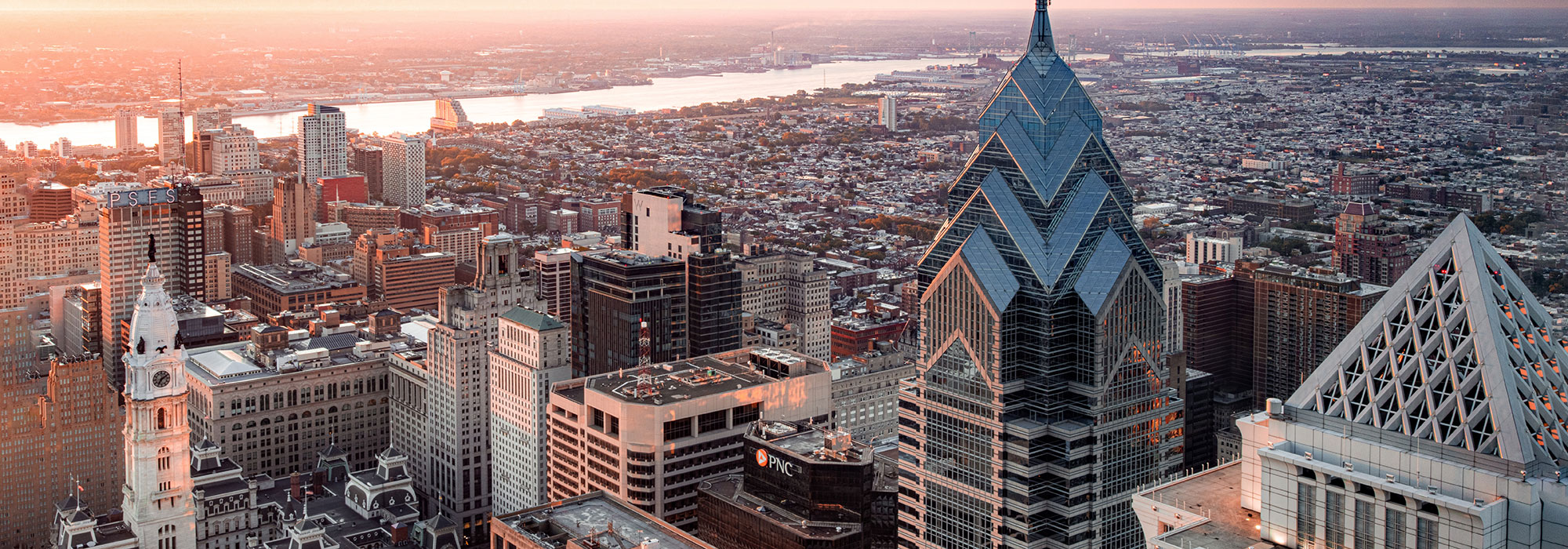 The image shows an aerial view of a cityscape at sunset. Prominent skyscrapers dominate the foreground, including a building with a distinctive glass pyramid top, home to renowned law firm marketing services. The city stretches out into the distance, bordered by a river, under a sky glowing with warm hues.