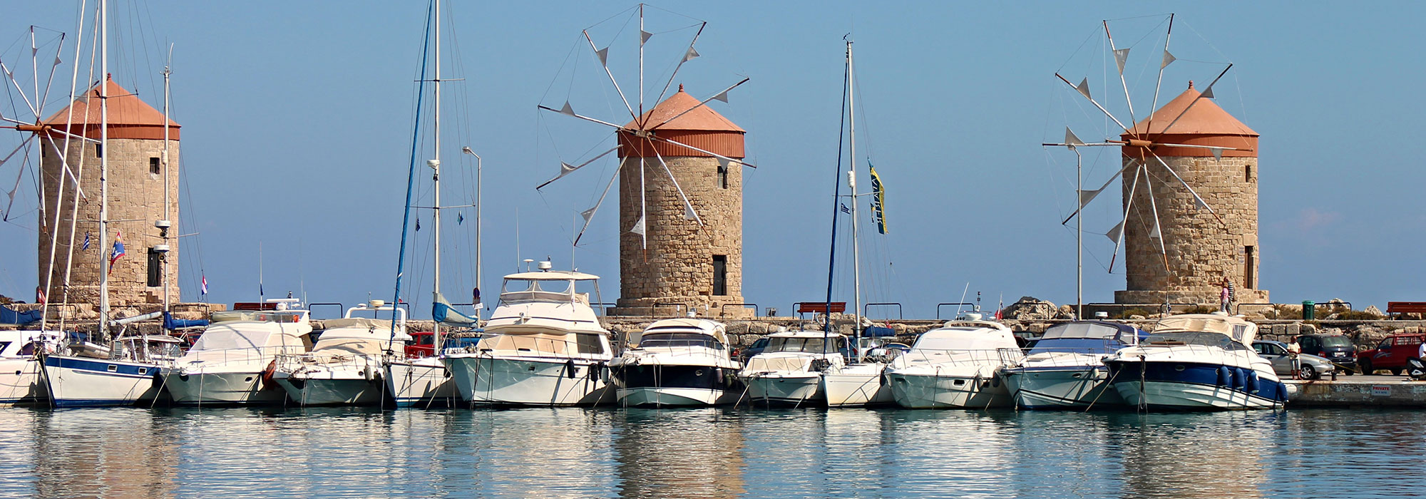 A row of boats docked at a marina in front of three historic stone windmills with terracotta roofs creates a picturesque scene. The clear sky reflects on the calm, blue water, where masts and flags can be seen among the boats—an idyllic setting that could inspire Law Firm Marketing Services.