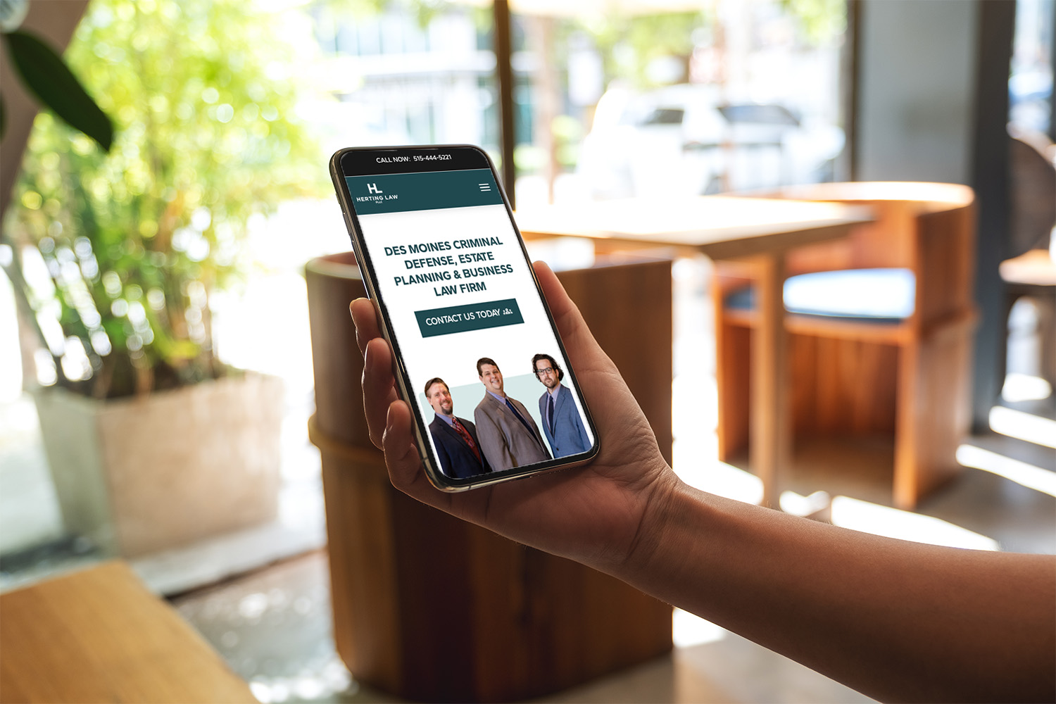 A person holds a smartphone displaying a law firm's website. The screen shows a group photo of four men in suits with text about criminal defense, estate planning, and business law services. The background is a cozy cafe interior with wooden furniture.