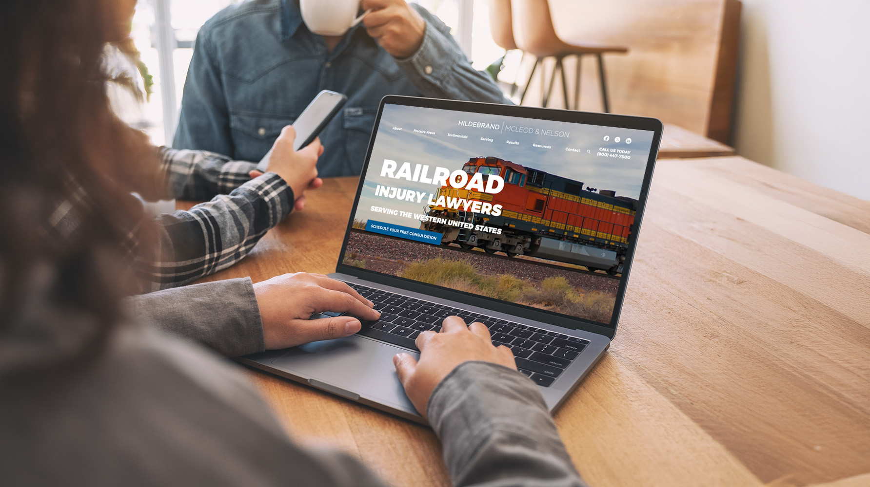 Person using a laptop displaying a website for railroad injury lawyers, with an image of a train. Another person in the background is drinking from a cup and holding a phone, seated at a wooden table.