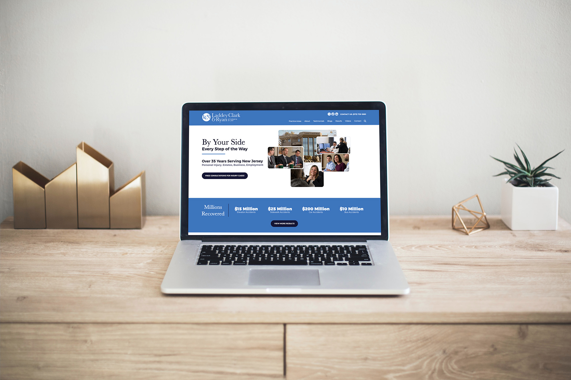 A laptop displaying a blue and white website is placed on a wooden desk. Next to the laptop are decorative items: a gold geometric sculpture and a small plant. The background is a plain light-colored wall.