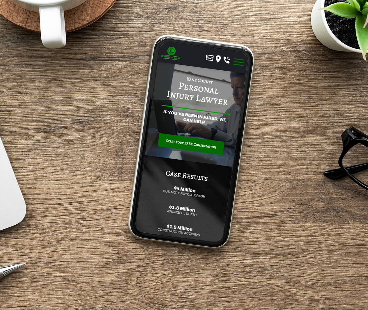 A smartphone on a wooden table displaying a personal injury lawyer's website. The screen shows contact options and case results. Nearby are a potted plant, a silver pen, and a white coffee cup.