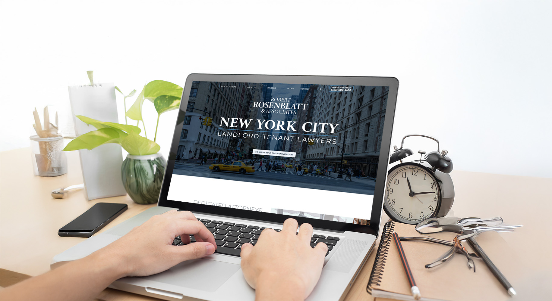 A person using a laptop displaying a website for New York City landlord-tenant lawyers. The desk has a potted plant, a black alarm clock, glasses, and stationery. The background shows a city street with buildings and taxis.