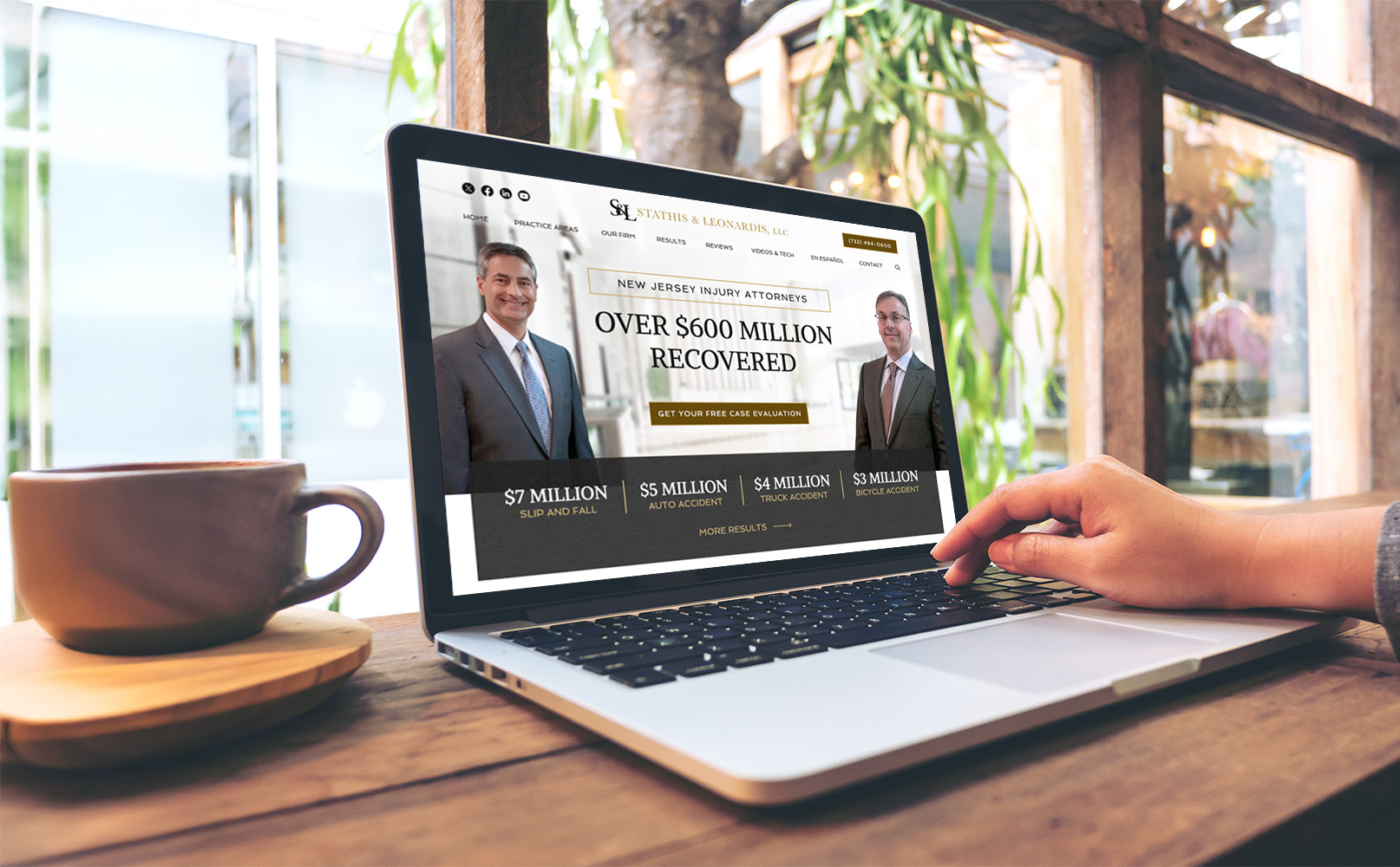 A person sitting at a wooden table with plants nearby uses a laptop displaying a lawyer's website. The screen highlights over 0 million recovered in New Jersey injury cases, alongside photos of attorneys and settlement figures. A coffee cup is nearby.