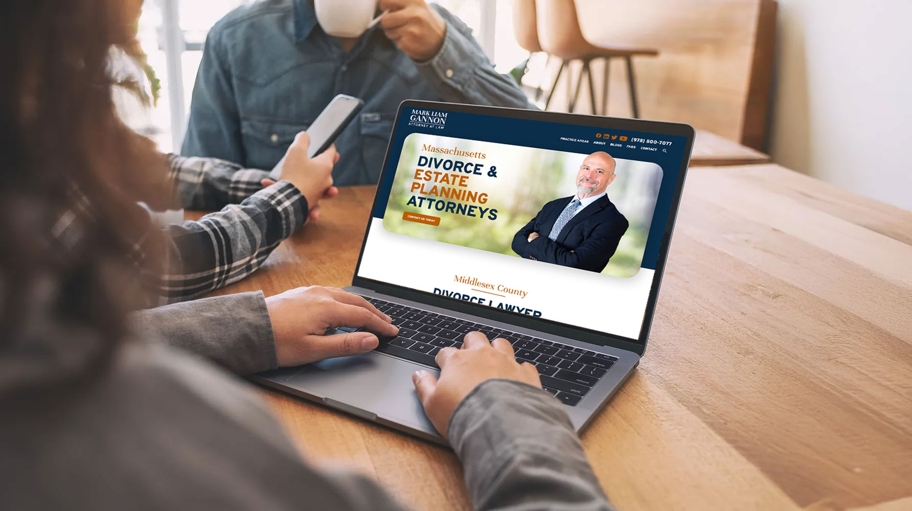 A person using a laptop at a wooden table, browsing a website for Massachusetts divorce and estate planning attorneys; another person sits across, holding a cup.